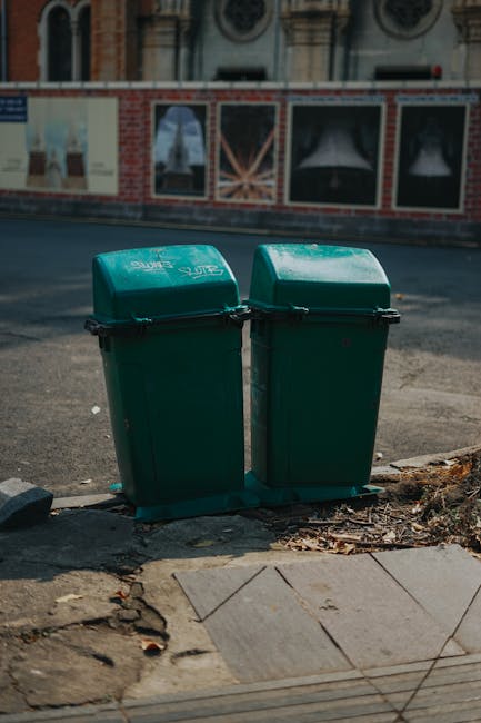A collection of discarded household waste and rubbish bags piled on the ground outdoors, positioned against a low stone wall. The waste includes black plastic garbage bags filled with debris, a yellow