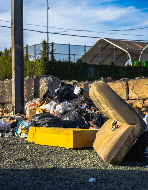 A sanitation worker dressed in a high-visibility yellow and orange vest operates a large red rubbish collection truck parked at the edge of a paved road. The worker appears to be managing waste inside