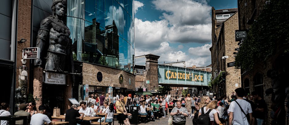 A busy outdoor scene at Camden Market in North London, featuring a diverse crowd of pedestrians seated at tables and walking along the street. In the foreground, various individuals enjoy their day, s