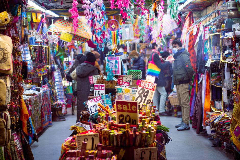 A busy outdoor scene at Camden Market in North London, featuring a diverse crowd of pedestrians seated at tables and walking along the street. In the foreground, various individuals enjoy their day, s