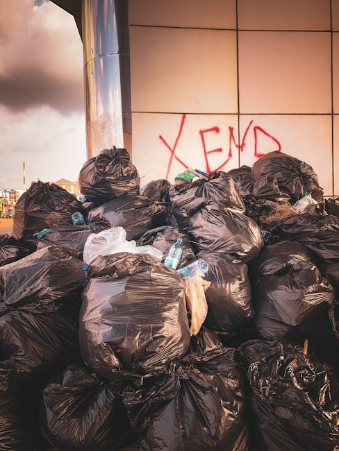 A large outdoor waste collection point in a paved urban area, featuring multiple black, red, and grey refuse bins filled with a mixture of household rubbish and paper waste. Several black plastic bags